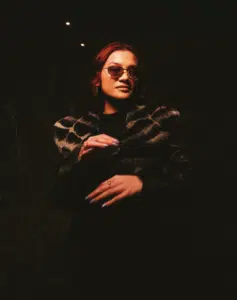 A person with sunglasses and a leopard-print fur jacket stands outdoors at night, dimly lit with hands raised in front of them.